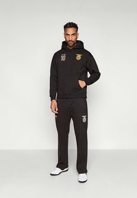 Homem vestindo um capuz preto e calças combinando com logótipos desportivos dourados e brancos, de pé com uma mão no bolso e ténis brancos.