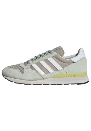 Jasnoszary i beżowy sneaker Adidas z białymi paskami, jasnozielonym akcentem na podeszwie i czarnym bieżnikiem, pokazany z profilu, skierowany w lewo.