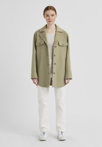 Olivenfarbene, übergroße Jacke mit großen Brusttaschen, runder Saum und Knopfverschluss, kombiniert mit weißen Hosen und weißen Turnschuhen.