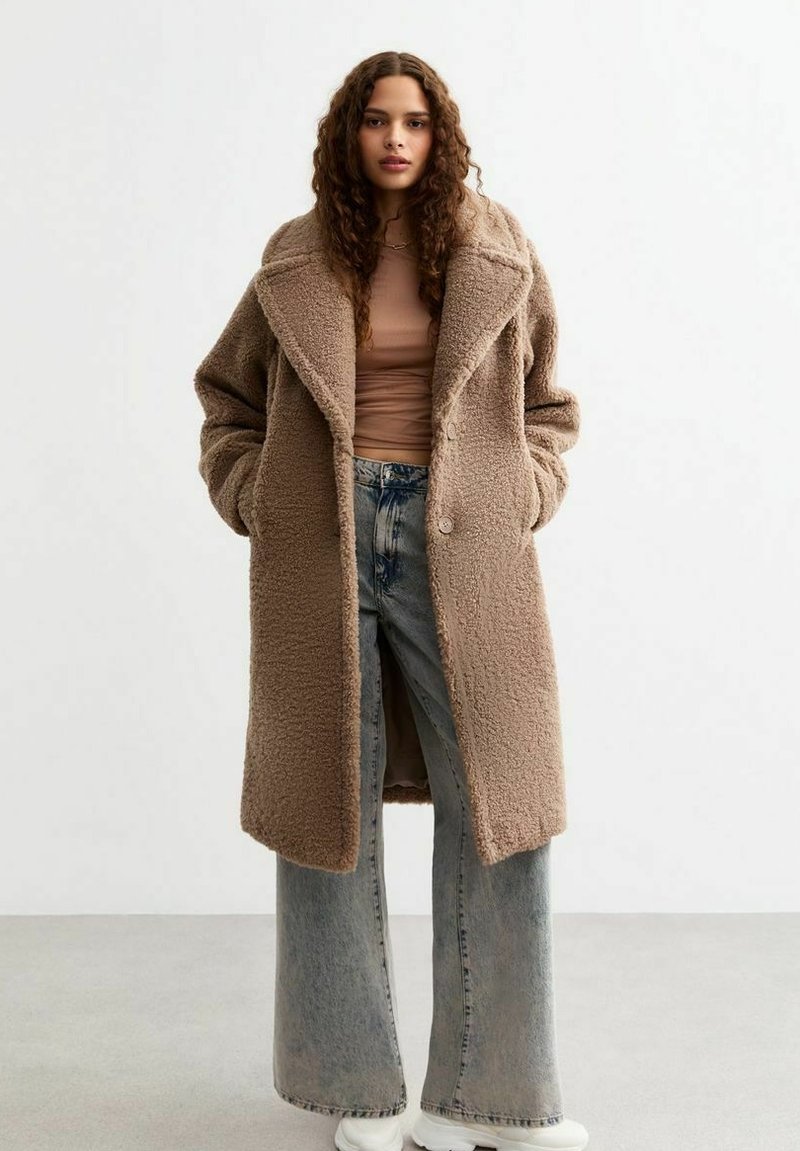Brun fleecejacka med stora slag, knapplåsning och sidofickor, lager på en ljusbeige topp och vida denimjeans.