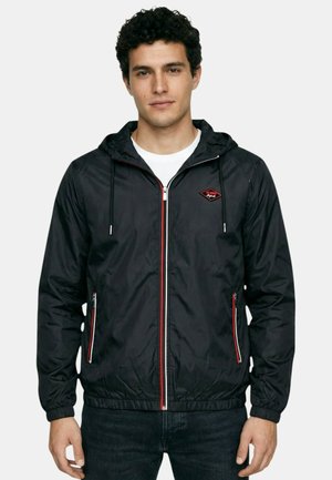 Jeune homme portant une veste à capuche noire zippée avec bordures rouges et blanches, debout les mains détendues le long du corps.