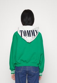 Sudadera verde con capucha blanca que presenta un gran logo azul de "TOMMY JEANS". Puños acanalados con rayas blancas y diseño de ajuste relajado.