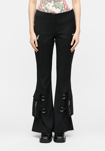 Μαύροι φαρδιοί παντελόνες cargo με πολλές τσέπες με φερμουάρ και αγκράφες, φορεμένοι με μαύρα παπούτσια με τετράγωνη μύτη.