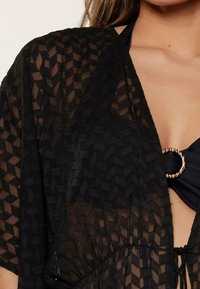 Kimono negro translúcido con un patrón en zigzag, que presenta mangas cortas y una cintura ajustable con cordón. Acento metálico circular en el escote.
