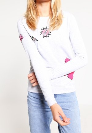 Frau trägt weißes Langarmshirt mit rosa Comic-Flicken und hellblaue Jeans, die Arme vor der Brust verschränkt.