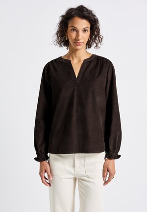 Woman wearing a long-sleeve dark brown blouse with a V-neck and ruffled cuffs, paired with high-waisted off-white pants.