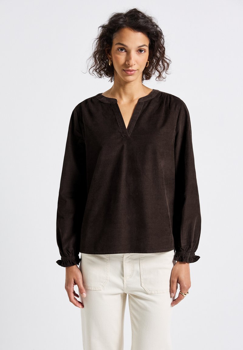 Vrouw in een donkerbruine blouse met lange mouwen, een V-hals en volants bij de mouwen, gecombineerd met hooggesneden offwhite broeken.