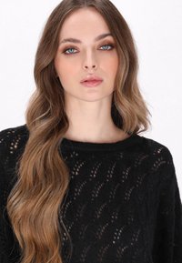 Maglione nero lavorato a maglia con una vestibilità ampia e un ampio scollo, caratterizzato da un motivo simile a pizzo e una finitura testurizzata. Capelli lisci e ondulati.