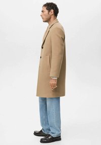 Abrigo largo de lana beige con diseño de un solo pecho, cuatro botones en el frente, bolsillos laterales, usado sobre un suéter ligero y combinado con vaqueros de mezclilla claros y zapatos negros.