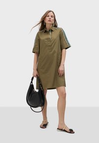 Olive green shirt dress with short sleeves, zipper collar, side pockets, and striped accents. Paired with a black handbag and floral flip-flops.
