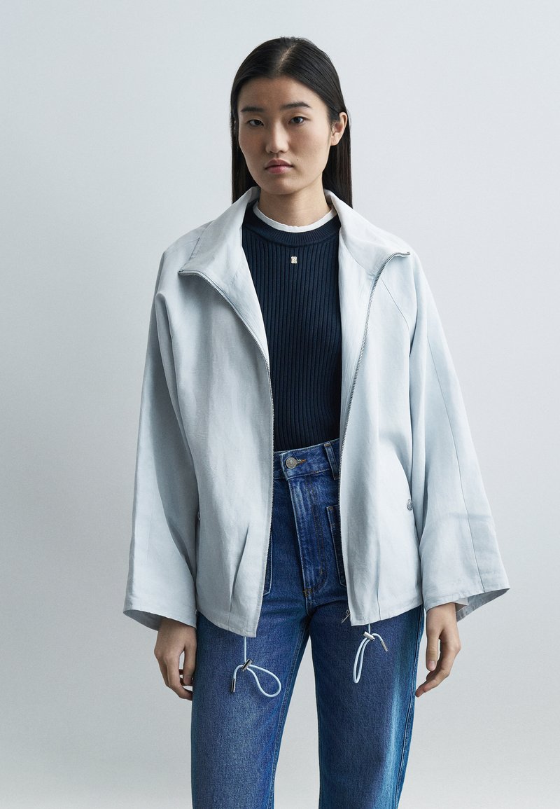 Νεαρή γυναίκα με μακριά σκούρα μαλλιά φοράει ένα ανοιχτόχρωμο, φαρδύ μπουφάν με φερμουάρ, μπλε ριμπ πλεκτή μπλούζα και ψηλόμεσα μπλε τζιν με κορδόνια.