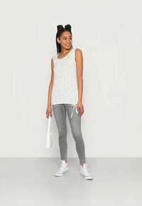 Top branco sem mangas com estampa floral pequena, combinado com jeans skinny cinza. A modelo usa ténis brancos e segura uma pequena bolsa branca.