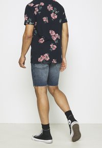 Lyhythihainen musta paita, jossa on vaaleanpunainen kukkakuosi, yhdistettynä haalistuneisiin denim-shortseihin ja mustiin korkeakärkisiin lenkkareihin. Rento ilme takaa.