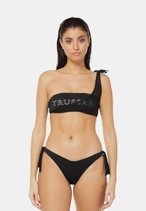Donna che indossa un top bikini nero monospalla con la scritta "TRUSSARDI" e slip bikini neri abbinati con laccetti laterali, in piedi su sfondo bianco.