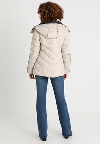 Beżowa kurtka wypełniona, z wzorem w zygzak, regulowanym kapturem i kontrastującymi szarymi akcentami. Noszona z niebieskimi dzwonami jeansami.