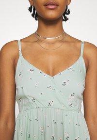 Ljusgrön blommig klänning med axelband, omslagsdesign och fina mönster av vita och orange blommor, tillverkad av mjukt tyg.