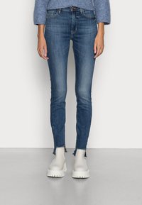 Jeans skinny azul oscuro con dobladillos deshilachados, combinados con botas blancas hasta el tobillo. La tela muestra un ligero desgaste, presentando un diseño clásico de cinco bolsillos.