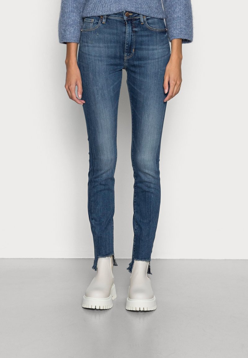 Jeans skinny azul oscuro con dobladillos deshilachados, combinados con botas blancas hasta el tobillo. La tela muestra un ligero desgaste, presentando un diseño clásico de cinco bolsillos.
