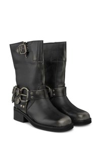 Bottoni in pelle nera alla caviglia con una punta lucida, fibbie decorative e una cintura con un anello metallico e rivetti sul lato.