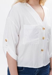 Weiße Bluse mit kurzen Ärmeln aus leichtem Stoff, mit V-Ausschnitt, zwei Brusttaschen und goldfarbenen Knöpfen auf der Vorderseite und den Ärmeln.