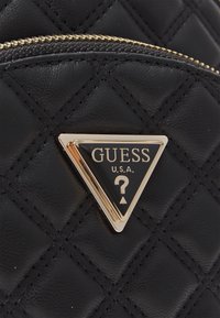 Schwarze gesteppte Handtasche mit einem auffälligen dreieckigen goldenen Logo-Schild, das eine schwarze und goldene Oberfläche sowie ein Fragezeichen aufweist. Reißverschlussdetails sichtbar.