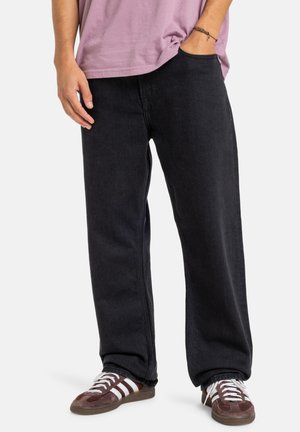 Pantalon en denim noir à coupe décontractée, avec des poches latérales et une jambe droite, associé à des chaussures de sport marron et blanches.