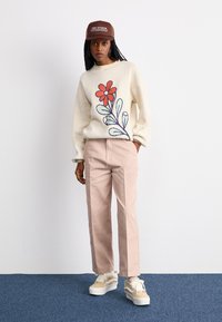 Cream sweater with red floral embroidery, light pink trousers, and brown cap; cream and tan sneakers with white laces. Textured materials.