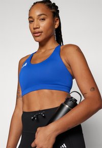 Blå sport bh med en tettsittende design, glatt tekstur og brede stropper, sammen med svarte høytalede leggings og en svart vannflaske.