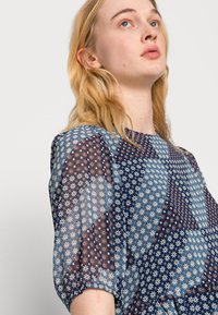Blusa transparente em tons de azul com padrões florais e designs geométricos. Mangas curtas bufantes com decote redondo e textura suave.