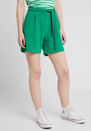 Person iført lysegrønne shorts med bælte, grøn- og hvidstribet top og hvide høje sneakers, stående på en lys grå gulv.