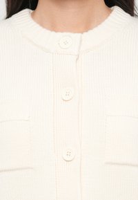 Kremfarget strikket cardigan med ribbestrikket krage, som har tre knapper og to påsydde lommer. Teksturen er myk og litt elastisk.
