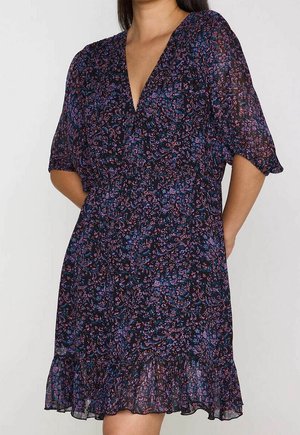 Vrouw die een donker bloemenjurk met V-hals, korte mouwen en een gerimpelde zoom draagt, staat met haar handen achter haar rug.