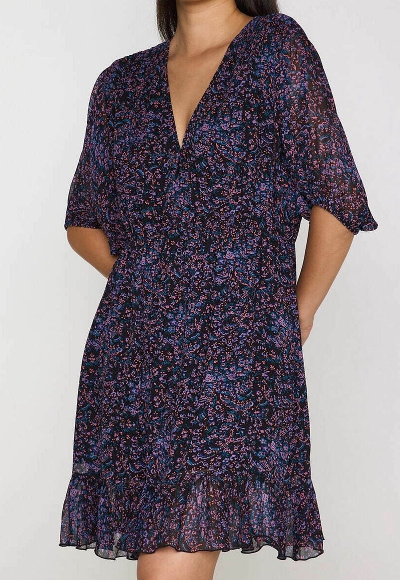 Femme portant une robe fleurie sombre à col en V, manches courtes et ourlet à volants, debout avec les mains dans le dos.