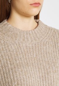 Beige stickad tröja med ribbad rund halsringning. Texturen är mjuk och tjock, med ett löst stickat mönster genom hela plagget.