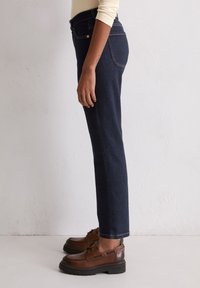Donkerblauwe rechte jeans met gouden stiksel, vijf zakken en een medium taille, gecombineerd met bruine leren schoenen en zwarte rubberen zolen.