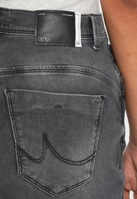 Stražnji pogled na sive traperice koje pokazuju šivenu unutrašnjost džepa, crni kožni brand patch s tekstom "l.t.b" i dio ruke osobe.