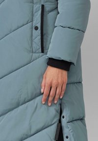 Gepolsterter Mantel in Hellblau mit Zickzacknaht, schwarzen Akzenten und einem seitlichen Reißverschluss. Verfügt über Rippbundabschlüsse und ein Markenlabel am Saum.