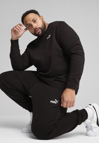 Felpa e pantaloni in cotone neri con logo PUMA bianco. Modello in ginocchio, indossando sneakers bianche. Texture liscia, vestibilità ampia.