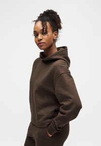 Νεαρή γυναίκα με σγουρά μαλλιά, ντυμένη με καφέ φούτερ και παντελόνι που ταιριάζει, στέκεται με το ένα χέρι στην τσέπη, στραμμένη ελαφρώς πλαγίως μπροστά σε ένα λευκό φόντο.