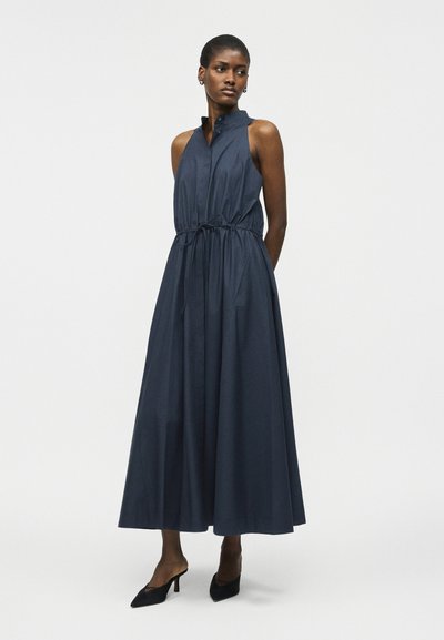Γυναίκα με αμάνικο, μακρύ σκούρο μπλε φόρεμα με ψηλό γιακά και ζωνάκι στη μέση, συνδυασμένο με μαύρα μυτερά τακούνια, στέκεται μπροστά σε λευκό φόντο.