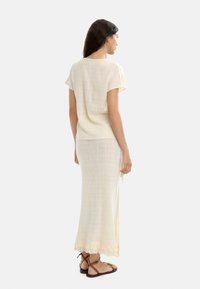 Jasno-beżowy zestaw składający się z bluzki z krótkim rękawem i długiej, prostej spódnicy z frędzlami u dołu. Wykonany z teksturowanej, lekkiej tkaniny.