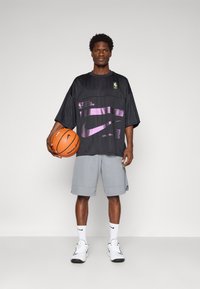 Black oversized jersey with purple accents and NBA branding, paired with gray shorts. Holds a basketball, wearing white socks and sneakers.