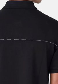 Polo noir avec un tissu texturé, présentant une teinte unie et une ligne horizontale contrastante à l'arrière avec de petits textes détaillés.