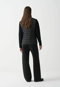 Schwarze Steppweste mit geometrischem Design, kombiniert mit einem schwarzen gestrickten Langarmshirt und eleganten schwarzen weit geschnittenen Hosen. Glatte Textur und taillierte Form.
