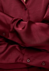 Camisa de seda burdeos con cuello abotonado, que presenta una textura suave, mangas largas y un brillo sutil. Los botones son redondos y del mismo color.