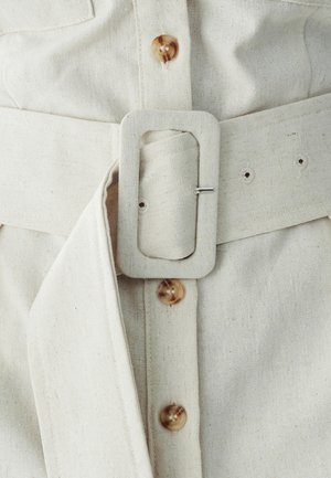 Crème linnen riem met een rechthoekige gesp, gevouwen stofuiteinde en bruine knoopdetails op een bijpassend kledingstuk. Gelaagd, eenvoudig ontwerp.
