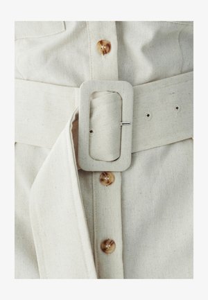 Krémový ľanový opasok s obdĺžnikovou prackou, prehnutým koncom látky a detailom hnedého gombíka na zodpovedajúcom odeve. Textúrovaný, jednoduchý dizajn.