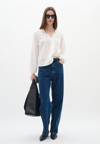 Camicia bianca a maniche lunghe con sottili righe verticali, jeans blu a vita alta e tacchi neri a punta. Borsa nera strutturata in mano.