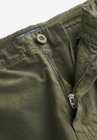 Pantaloni dintr-o țesătură ușoară, de culoare verde măsliniu, cu fermoar vizibil la brâu, închidere cu nasture și talie texturată. Prezintă o etichetă pe spate.
