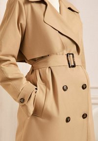 Trench coat bej cu un guler larg, nasturi dubli, curea cu cataramă pătrată și buzunare laterale. Material neted, texturat.